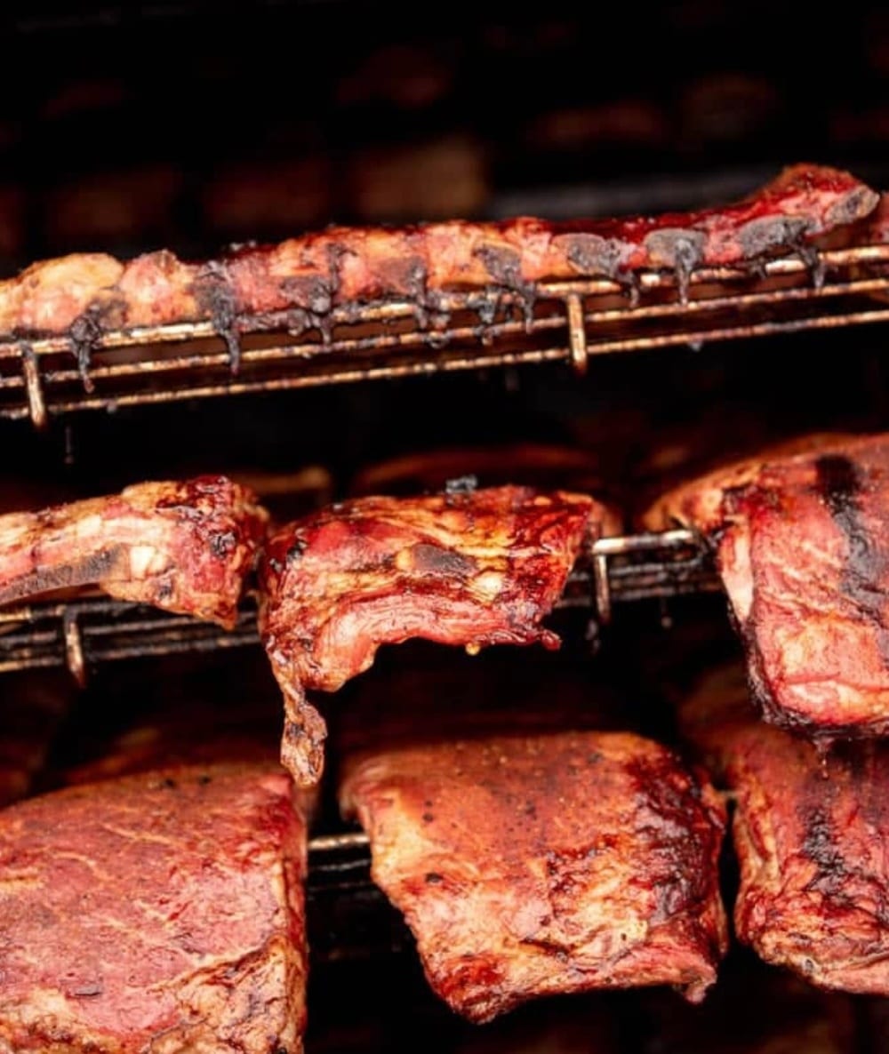 Racks of glazed pork ribs cooking on the wire shelves of a smoker, the bark dark and the edges caramelised.
