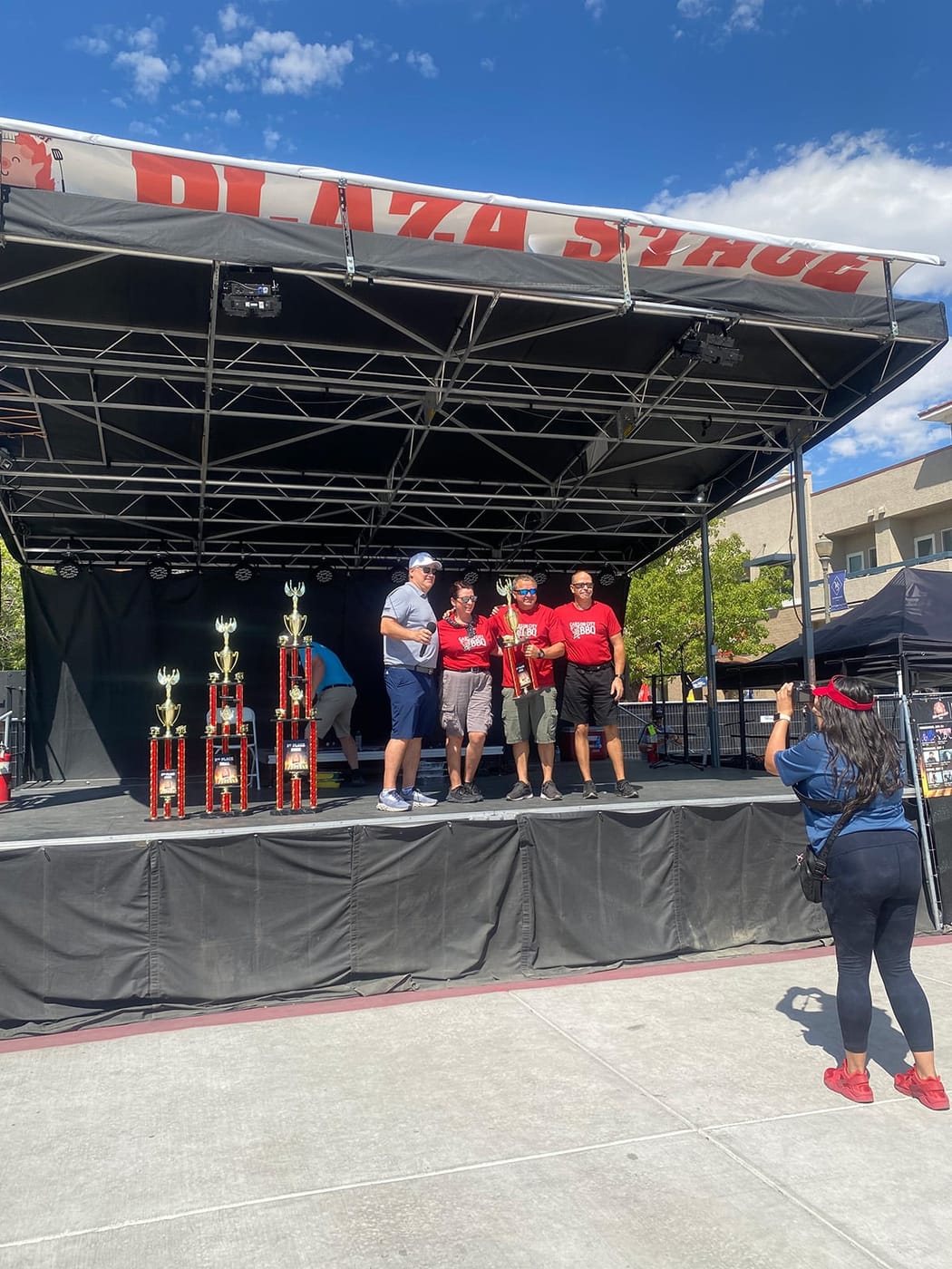 Carson City BBQ team members standing on an outdoor cook-off stage beside three tall trophies during the awards presentation.