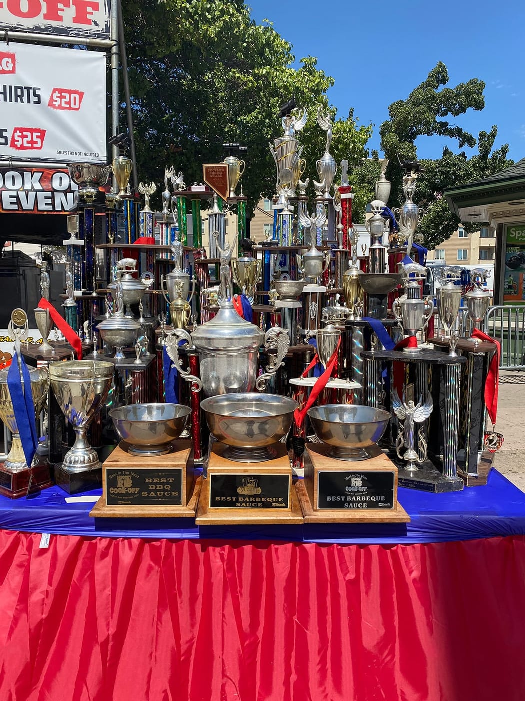 A massed display of silver cups, trophies and ribbons arranged on a red and blue draped table at a cook-off booth.