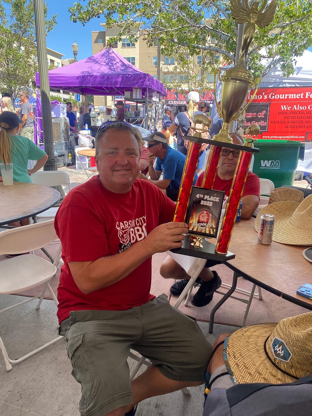 A man in a red Carson City BBQ shirt seated at an event table holding up a fourth-place cook-off trophy.