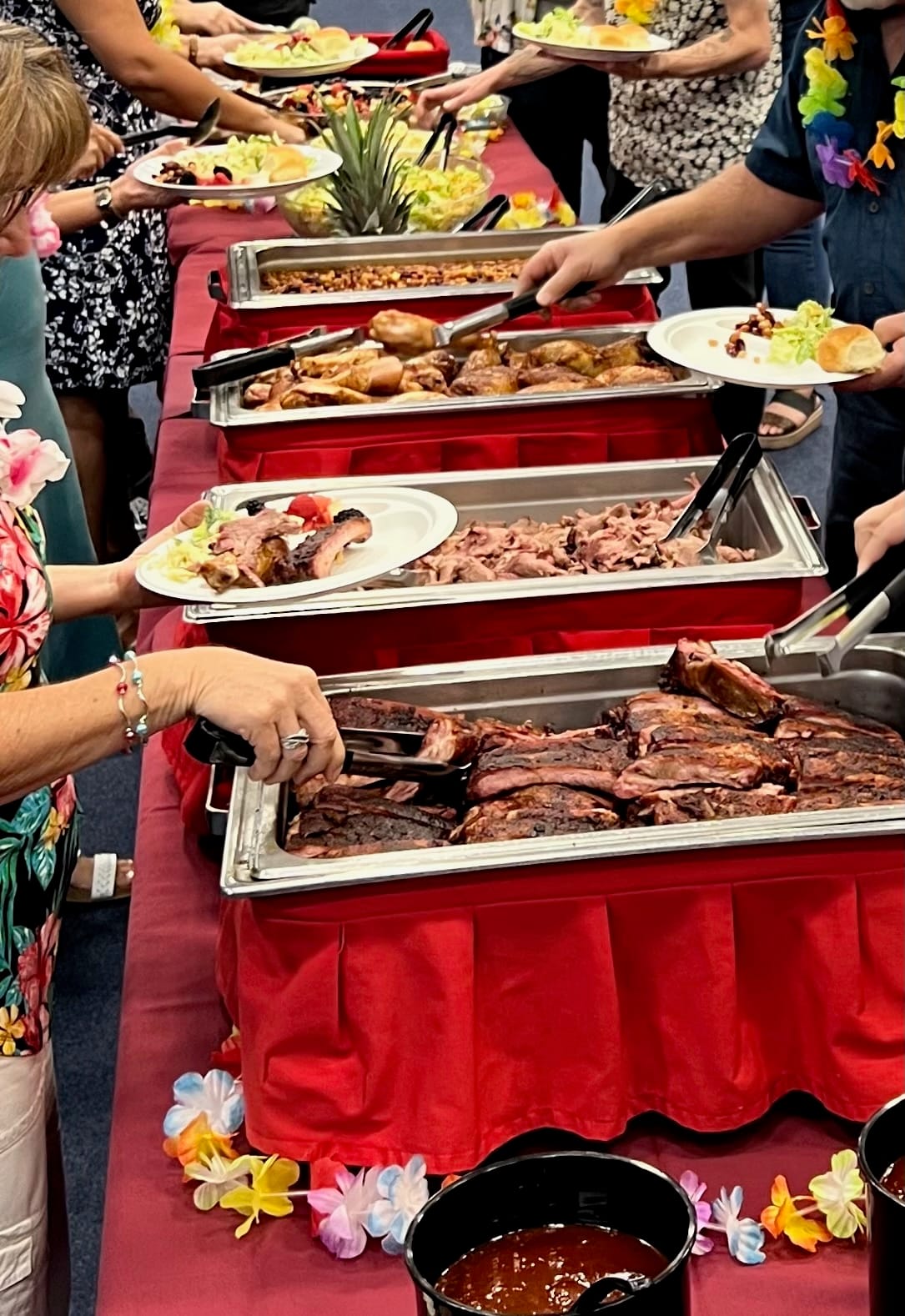 Guests filling their plates along a Carson City BBQ buffet line: chafing trays of ribs, pulled pork, chicken and baked beans on a red-skirted table.