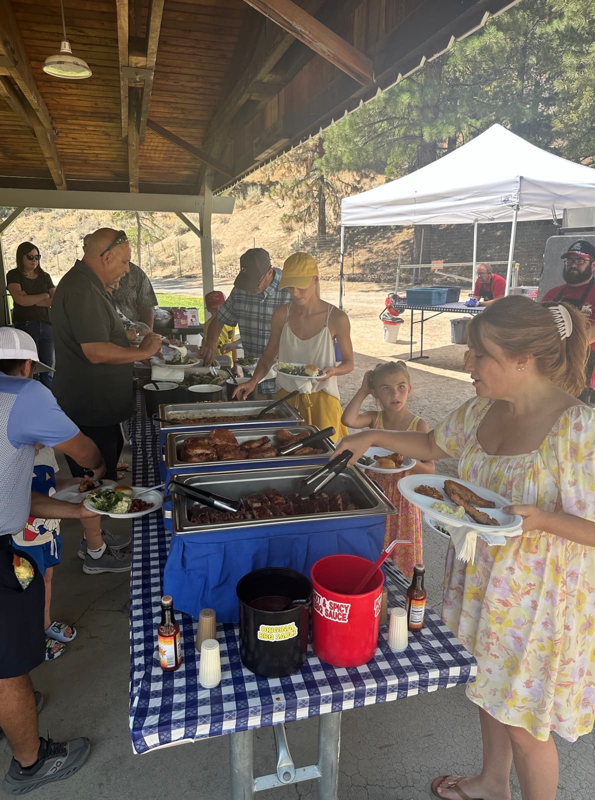 Families filing along a Carson City BBQ buffet line under a park shelter, filling plates from chafing trays of chicken and ribs on a blue-checked tablecloth.