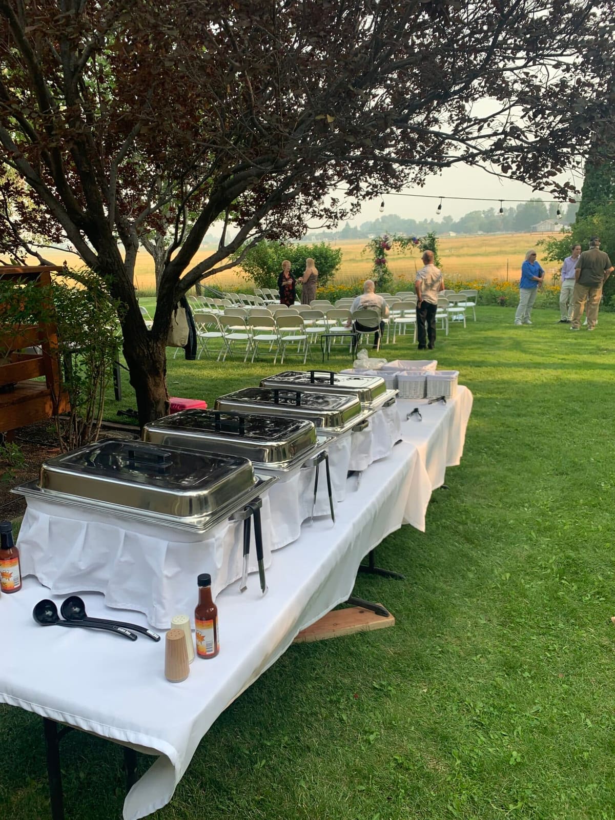 Chafing dishes and a bottle of sauce along a white-clothed buffet table on a lawn beneath a tree, rows of folding chairs set out for a ceremony and open pasture beyond.