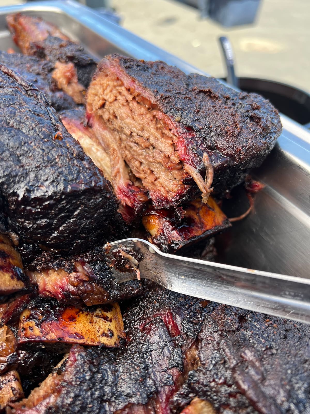 Tongs lifting a portion from a tray of dark, charred-edged smoked meat.