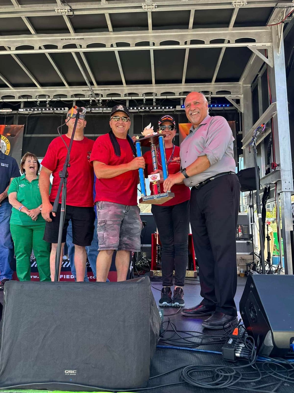 Carson City BBQ team members standing on an outdoor stage accepting a trophy from an event official in a dress shirt and tie.