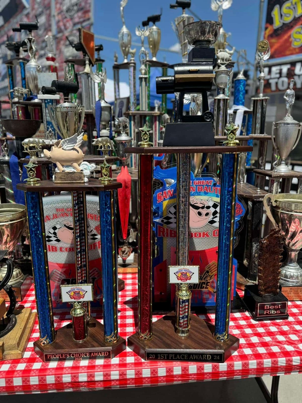 A row of tall competition trophies with checkered-flag plaques standing on a red-and-white checked tablecloth at the Carson City BBQ booth.