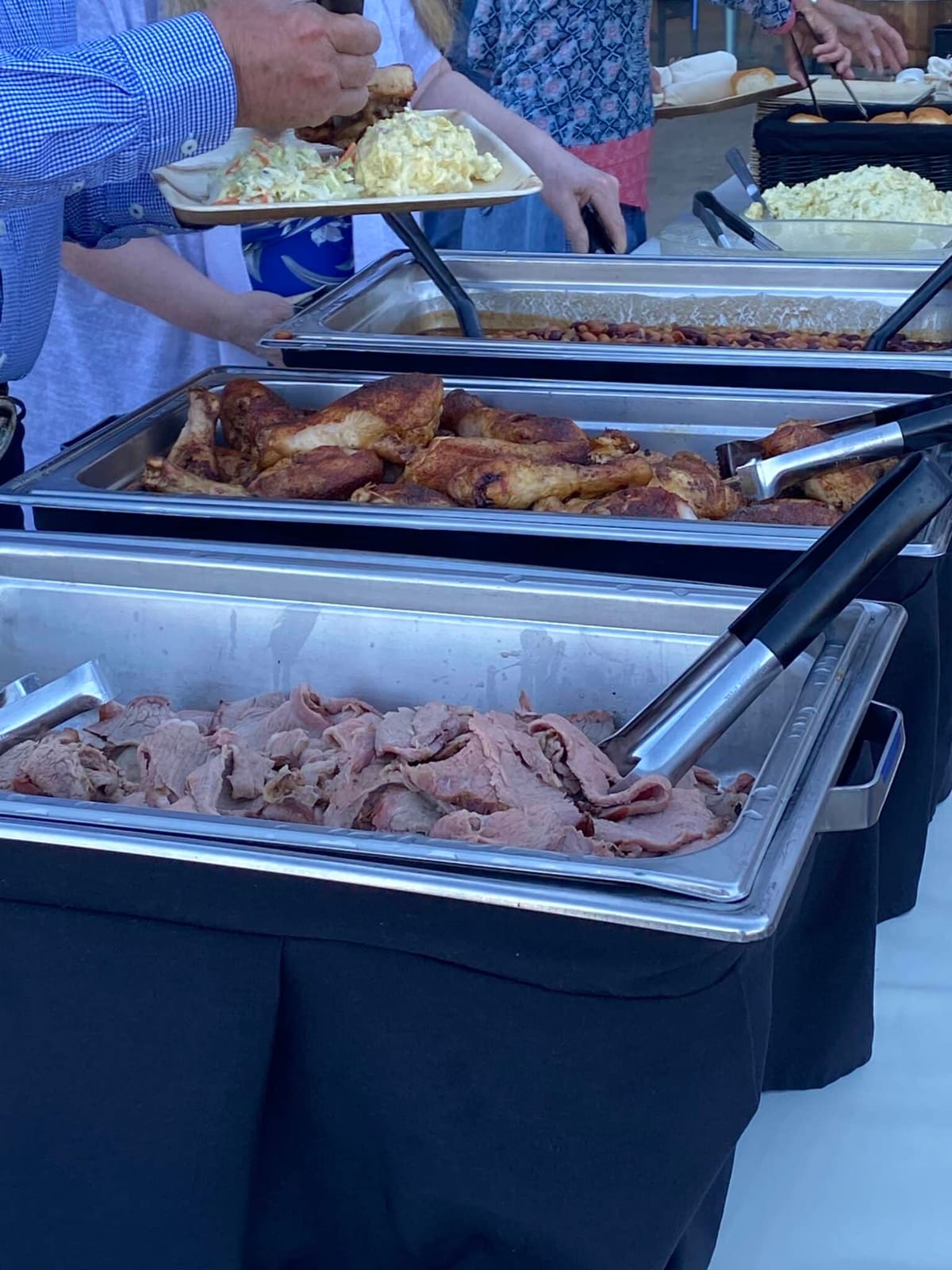 Guests filling paper plates from a line of chafing trays holding roast chicken, sliced beef, beans and coleslaw, with a basket of rolls at the far end of the table.