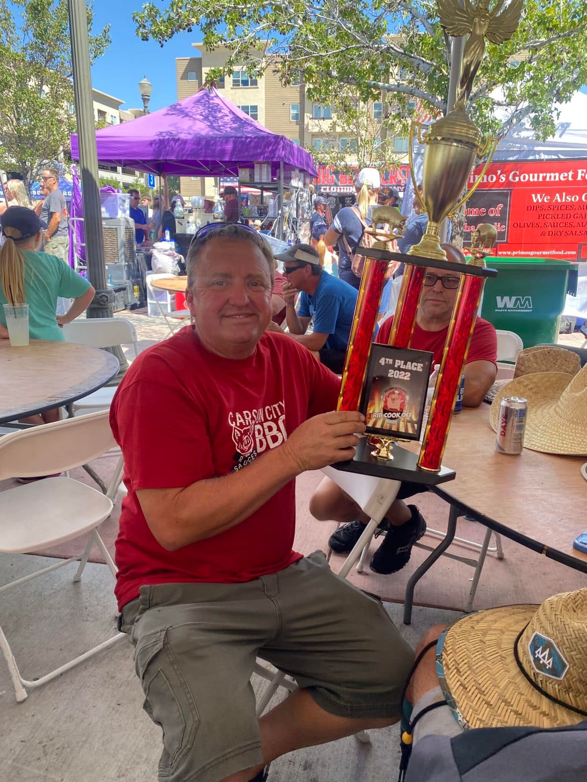 A man in a red Carson City BBQ shirt sitting at an event table holding up a trophy toward the camera.