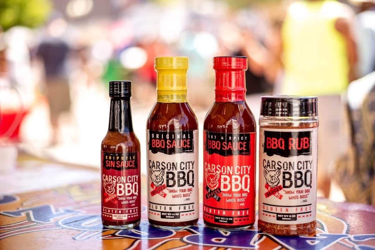 All four Carson City BBQ products lined up on a brightly tiled table at an outdoor event with a blurred crowd behind them: the tall black-capped Chipotle Sin Sauce, the yellow-capped Original BBQ Sauce, the red-capped Hot & Spicy BBQ Sauce, and the BBQ Rub shaker.