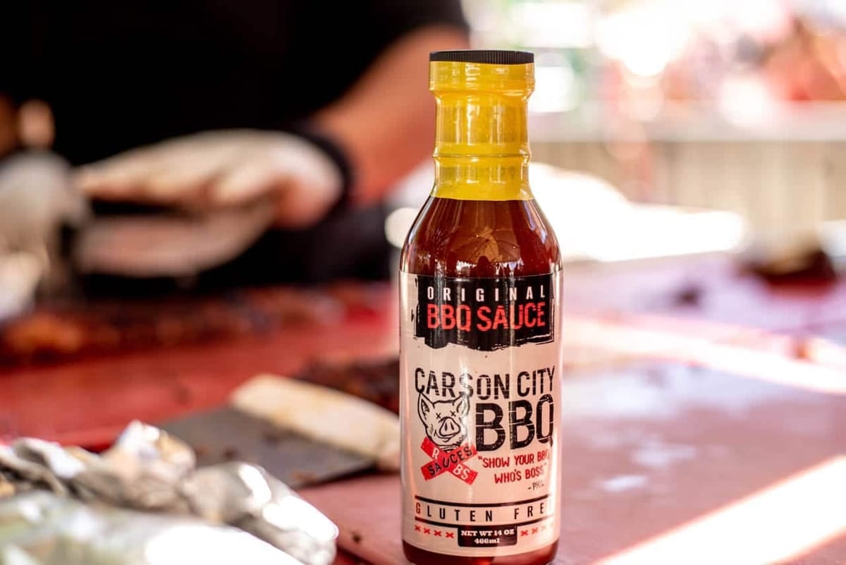 A bottle of Carson City BBQ Original BBQ Sauce with a yellow cap standing on a table at an outdoor cook, a gloved cook working at the prep table behind it