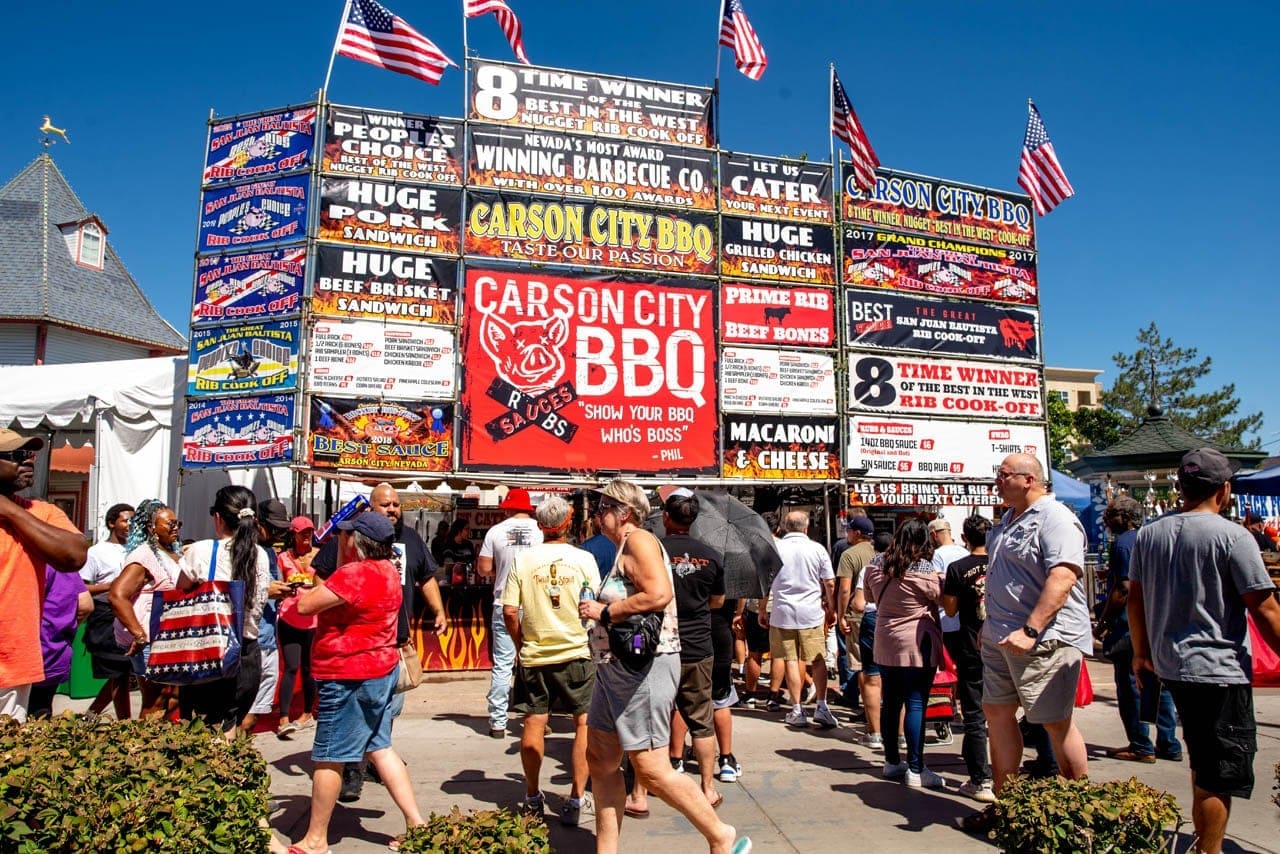 Festival crowds walking beneath the towering Carson City BBQ cook-off booth signs, which advertise ribs, brisket, macaroni and cheese and a "Show your BBQ who's boss" pig logo.