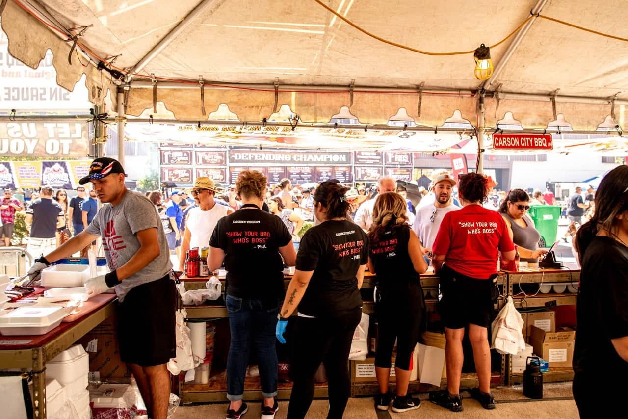 The Carson City BBQ crew working shoulder to shoulder along the service counter inside their tent, plating orders while the festival crowd passes outside