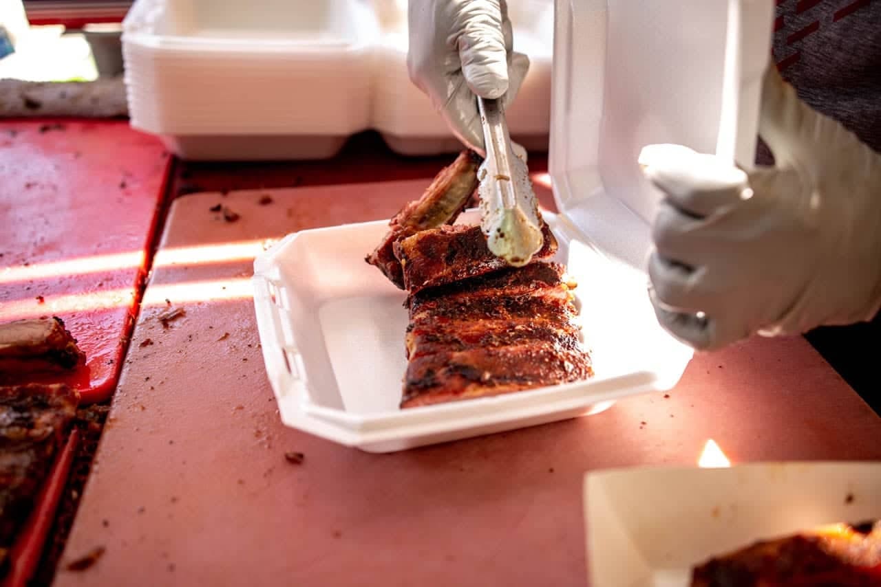 A gloved hand using tongs to lift a portion of glazed ribs into a white takeout clamshell on a red cutting board.