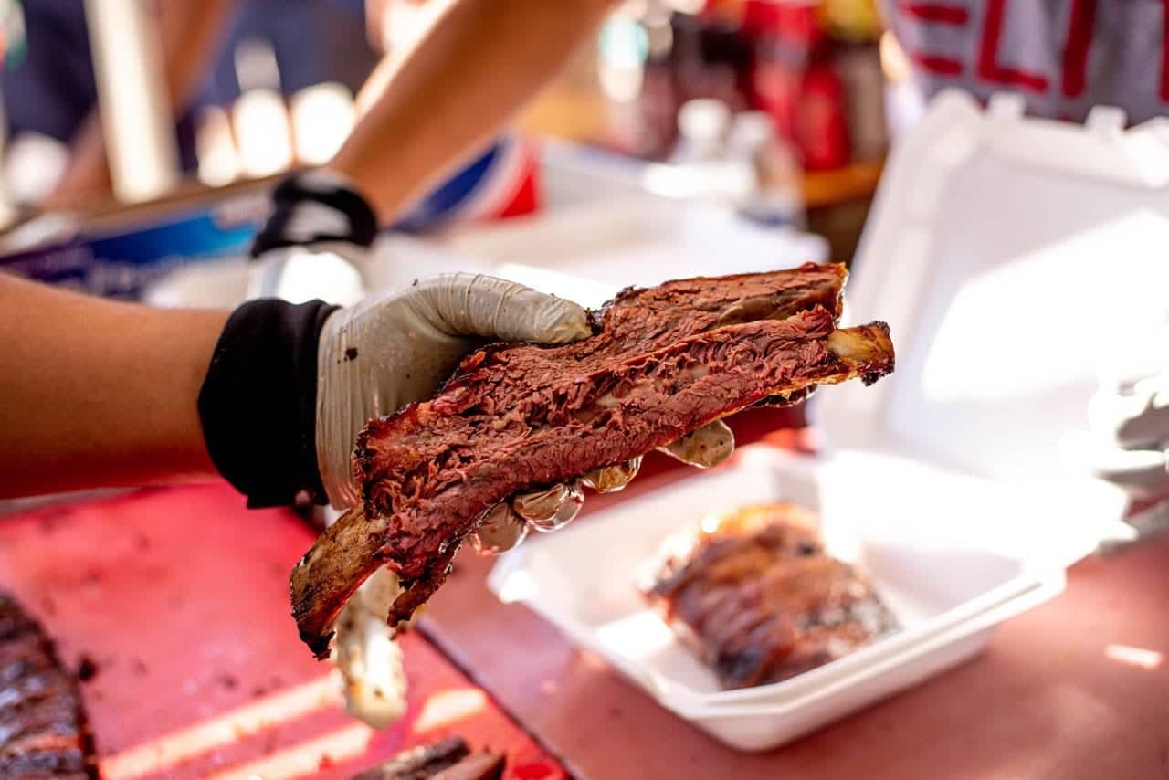 Gloved hands holding up a dark-crusted rack of ribs above a red prep table, with a filled takeout container waiting beside it.