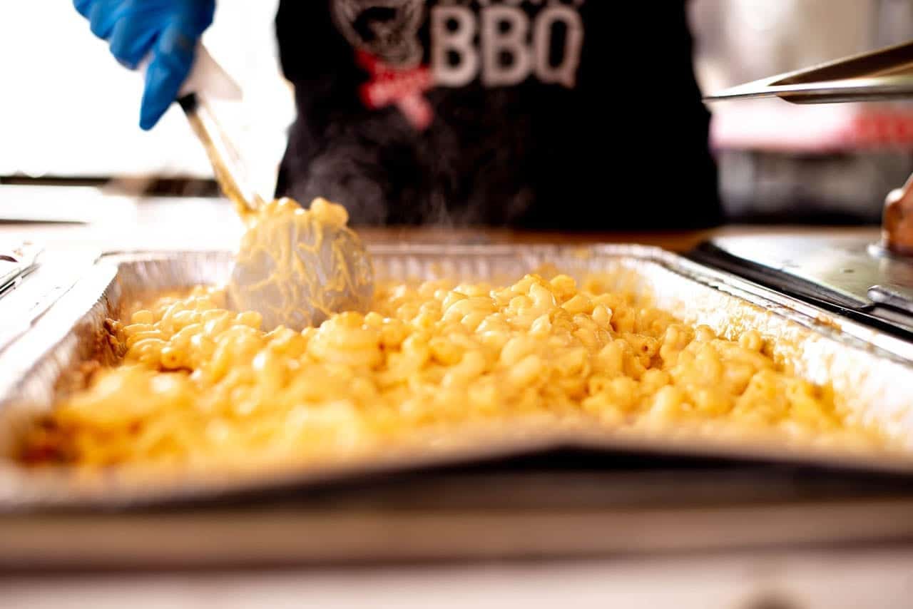 A crew member in a Carson City BBQ shirt scooping macaroni and cheese from a full steam-table tray.