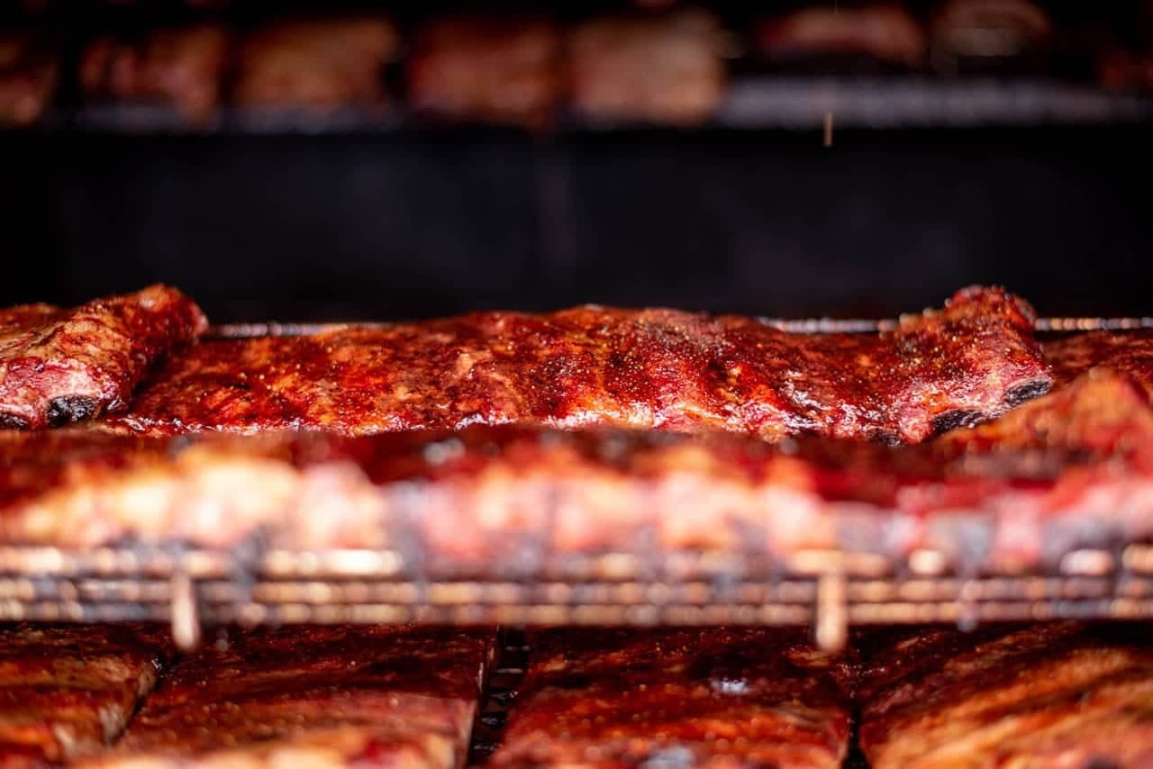 Long rows of ribs stacked on the racks inside a rotisserie smoker, their surfaces darkened and lacquered from the heat.