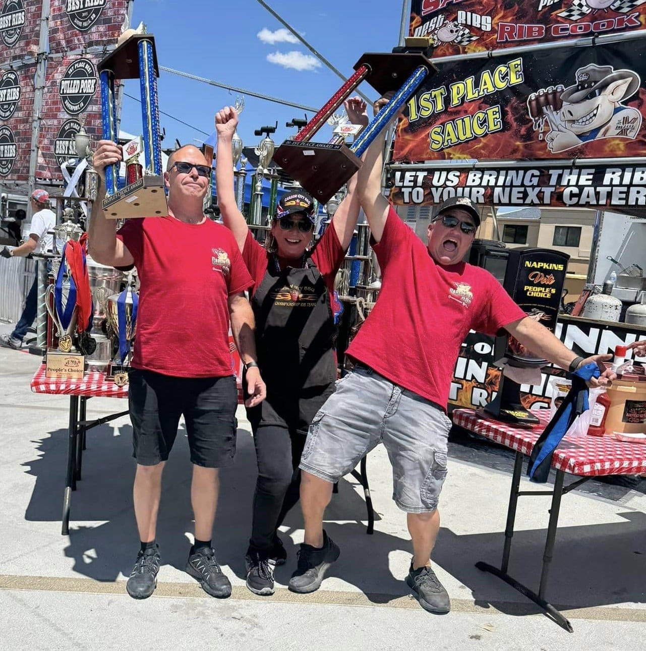 Three Carson City BBQ crew in red team shirts celebrating at a rib cook-off, two of them holding first-place trophies overhead in front of a 1st Place Sauce banner.
