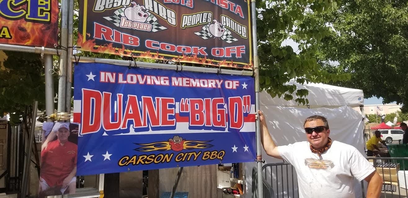A red, white and blue banner reading “In loving memory of Duane ‘Big D’ — Carson City BBQ”, hung on the team's booth at the San Juan Bautista Rib Cook Off beneath their first-place Best Ribs and People's Choice banner, a photograph of Duane Felker in his chef's coat fixed at one end.