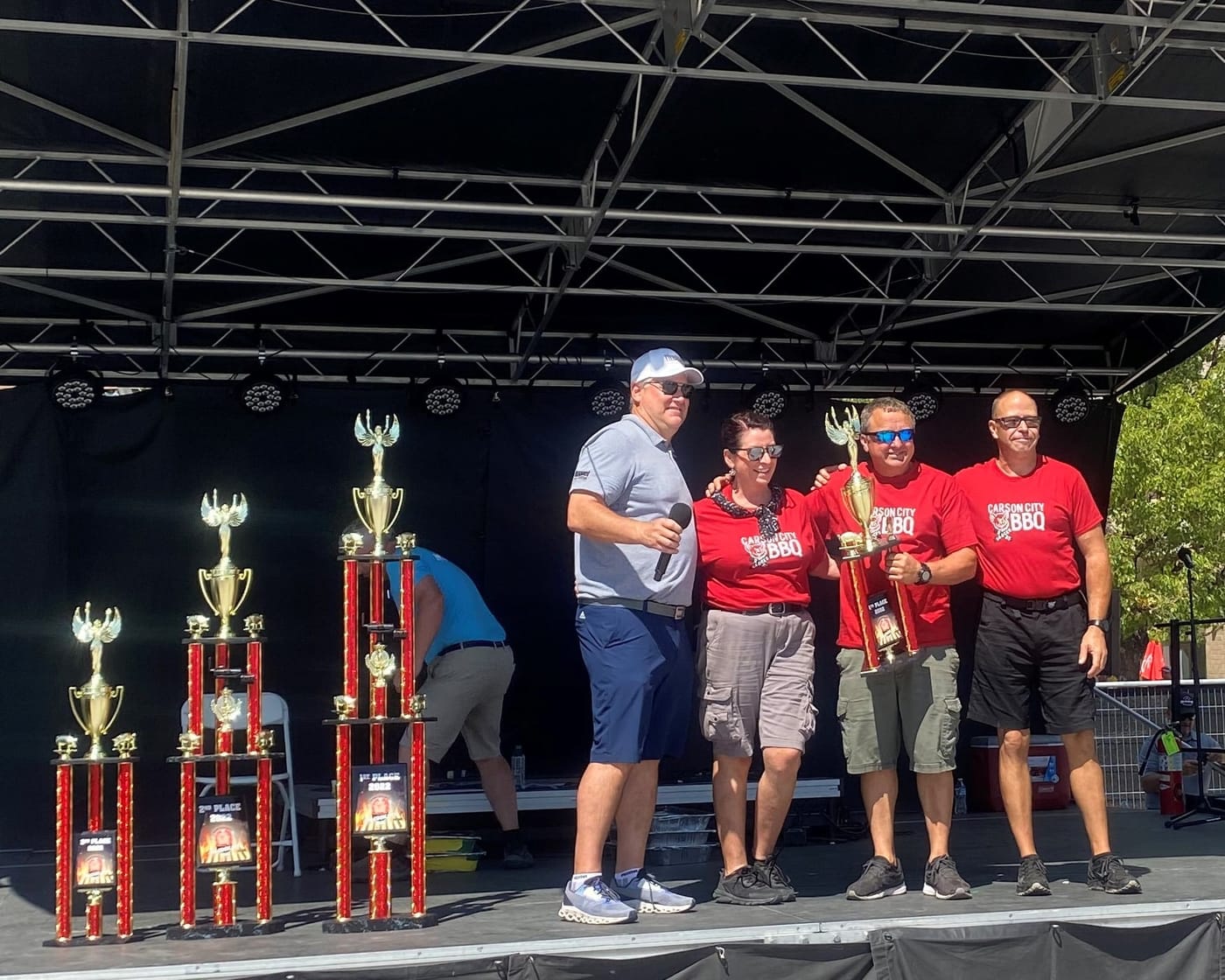 Carson City BBQ on the winners' stage at a rib cook-off — three team members in red Carson City BBQ shirts, one holding a first-place trophy, standing with the event's host, three tall trophies lined up beside them.