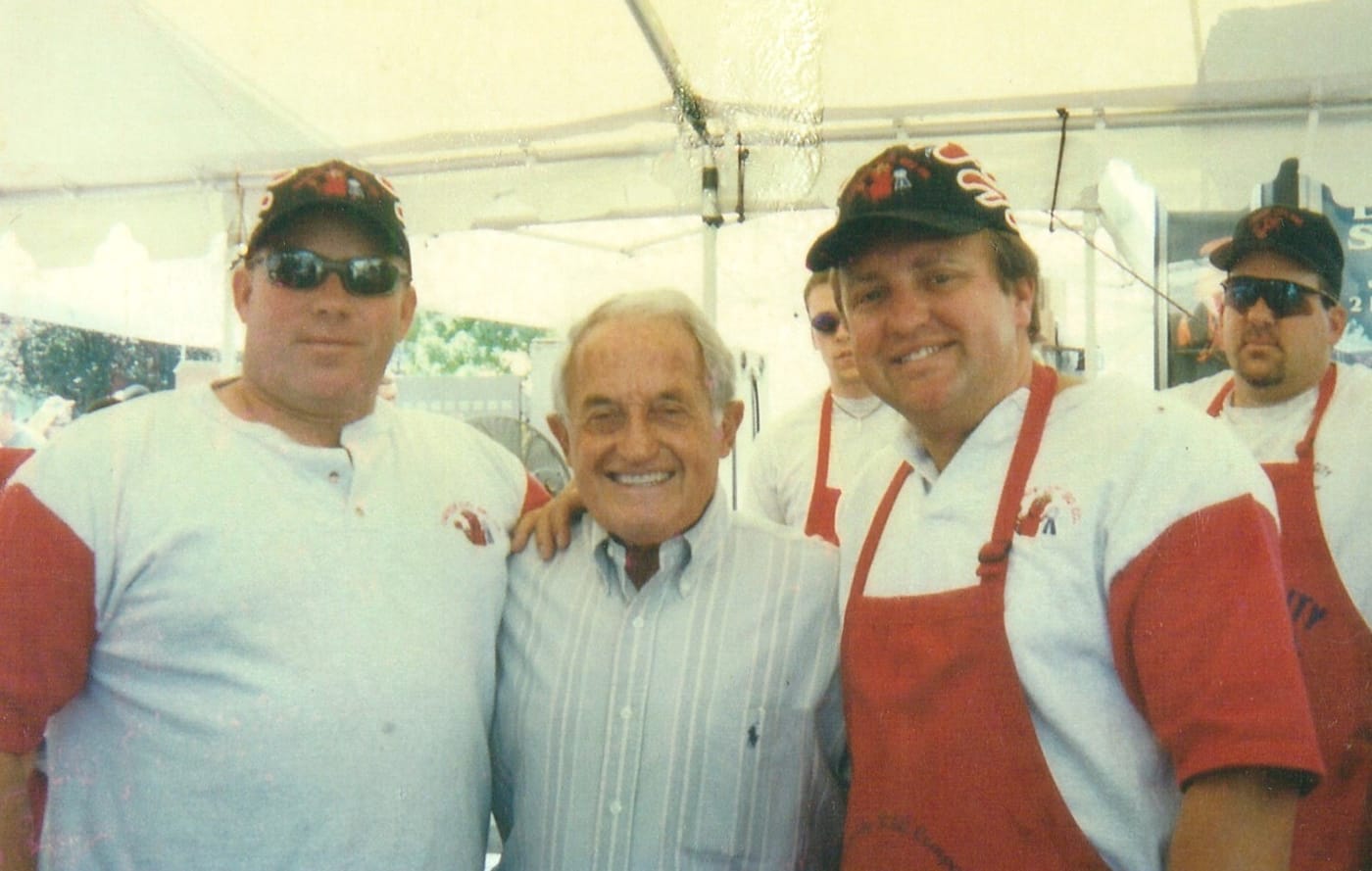 Phil Hyatt, Duane Felker and John Ascuaga, founder of John Ascuaga's Nugget, smiling together under a white cook-off tent — two in matching red-and-white Carson City BBQ shirts and caps, one in a striped button-down shirt — with more of the crew in red aprons behind them.