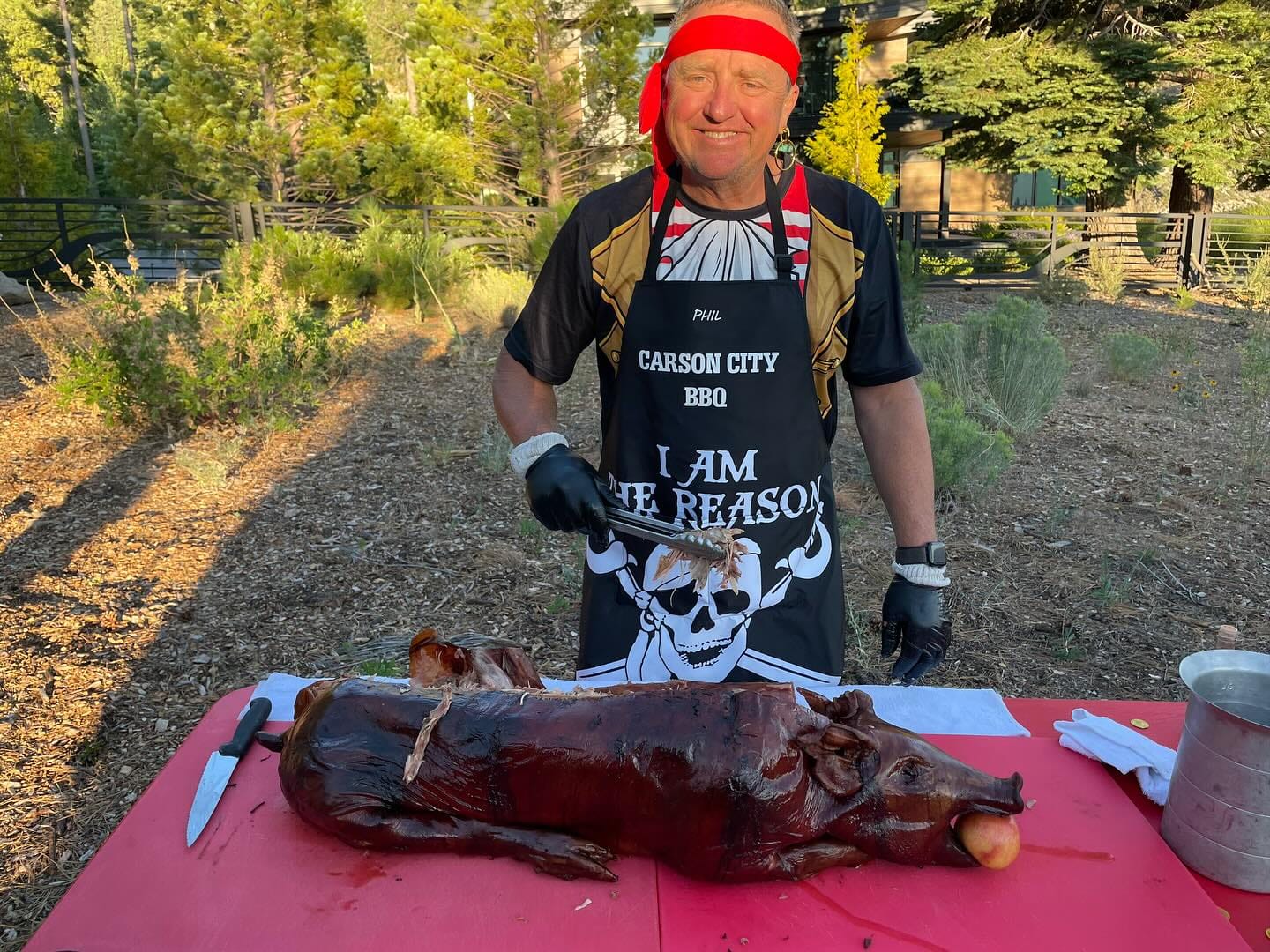A cook in a Carson City BBQ apron and a red headscarf standing over a whole roasted hog on a red cutting board at a table set up on open sagebrush ground in the evening light.