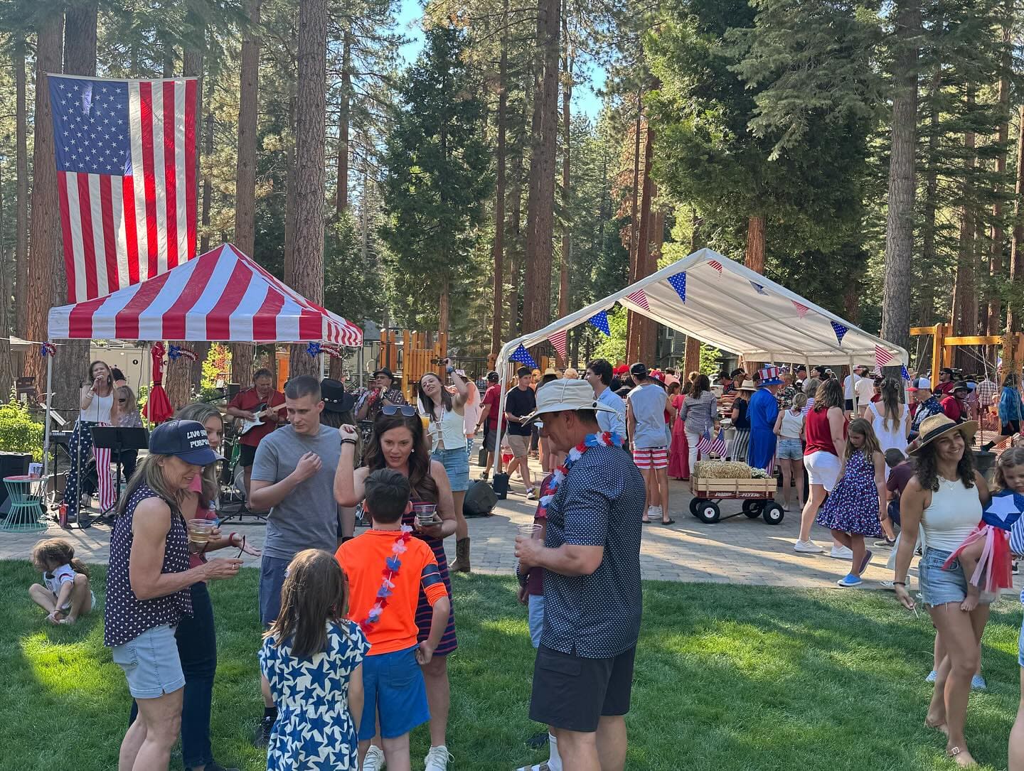 Guests standing with drinks on a lawn between two canopies at an outdoor summer party, a band playing under tall pines behind them.