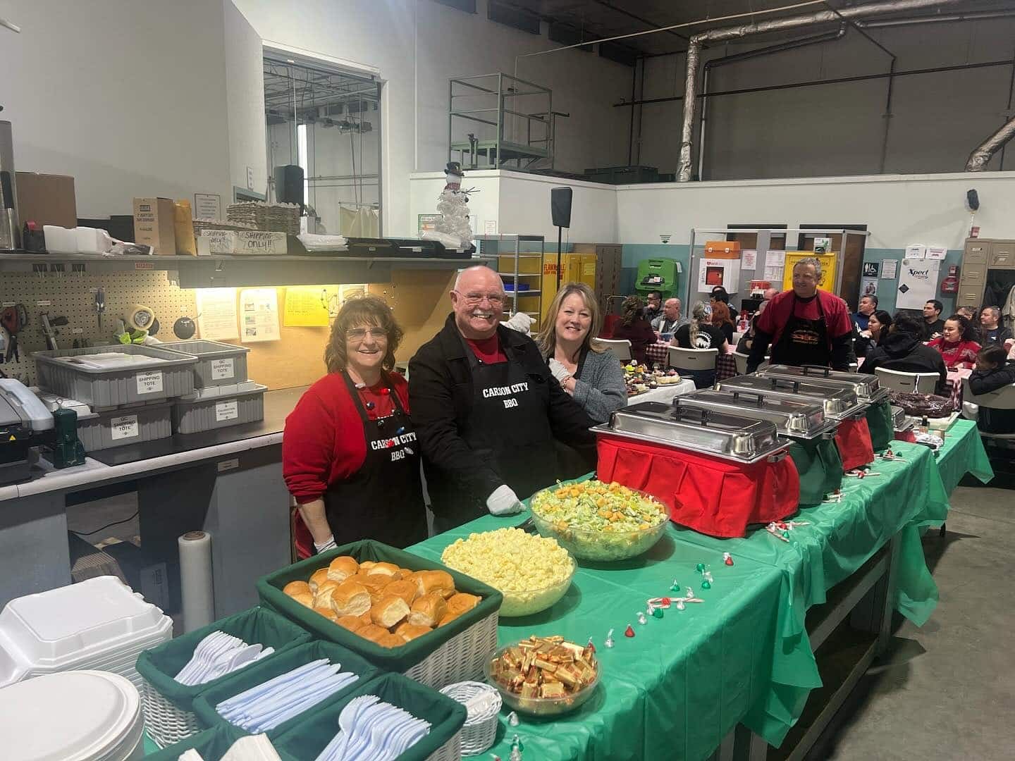 Two people in Carson City BBQ aprons standing behind a long green-clothed serving table of rolls, salads and chafing dishes set up inside a working warehouse, with staff eating at tables behind them.