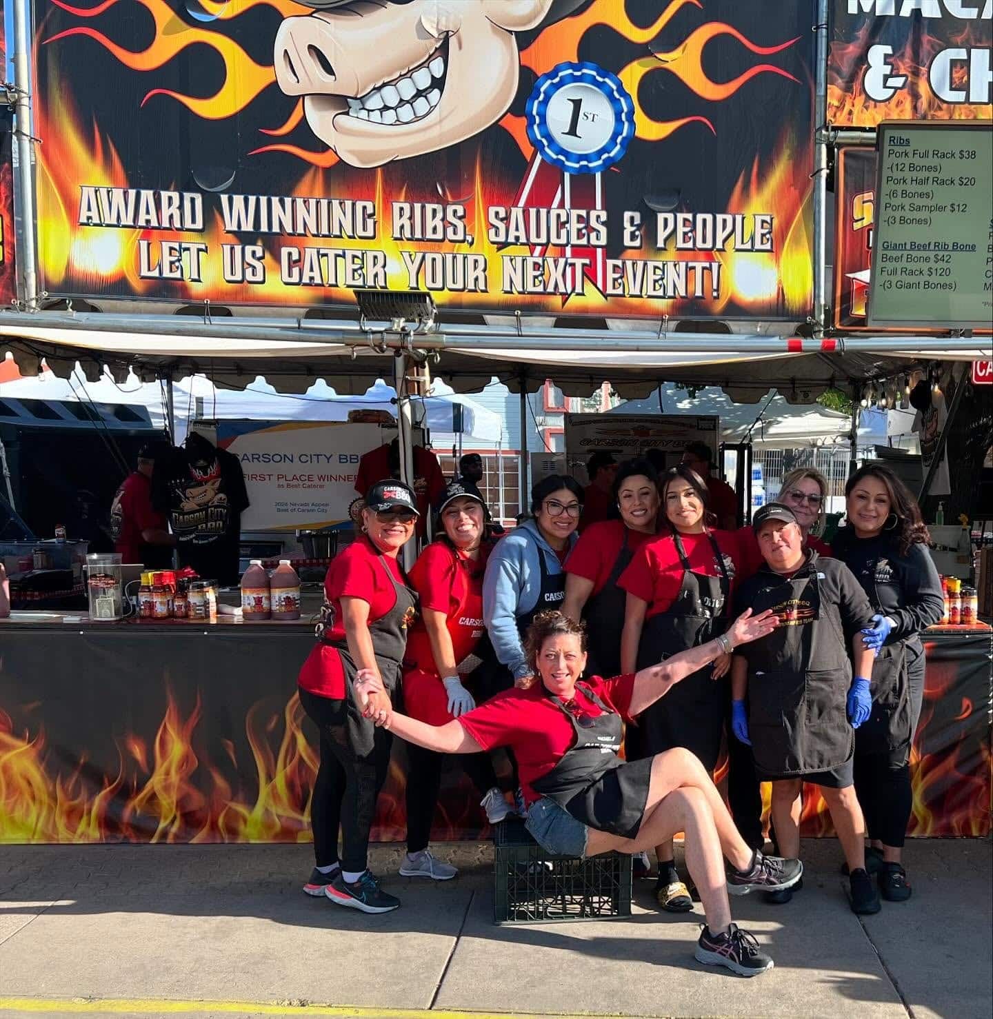 About ten Carson City BBQ team members in red and black shirts posing together in front of the flame-painted booth front, which reads "Award winning ribs, sauces & people — let us cater your next event!".