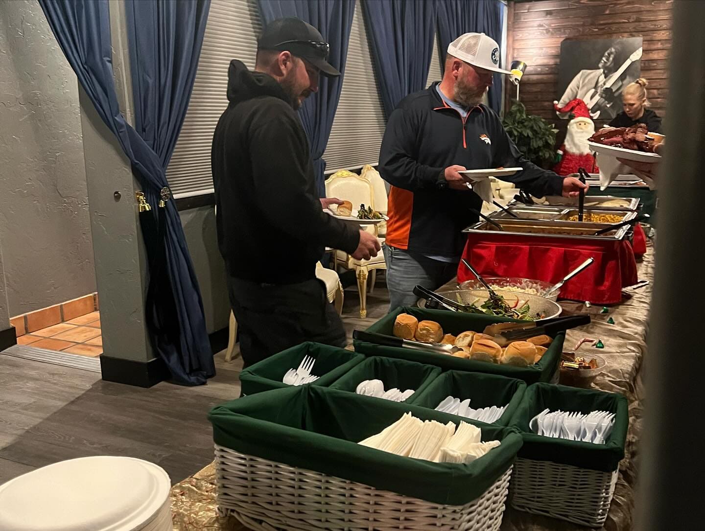 Guests in jackets and caps filling plates along an indoor buffet of chafing trays, rolls and salad, with cutlery and napkin baskets in the foreground and a decorated room behind.