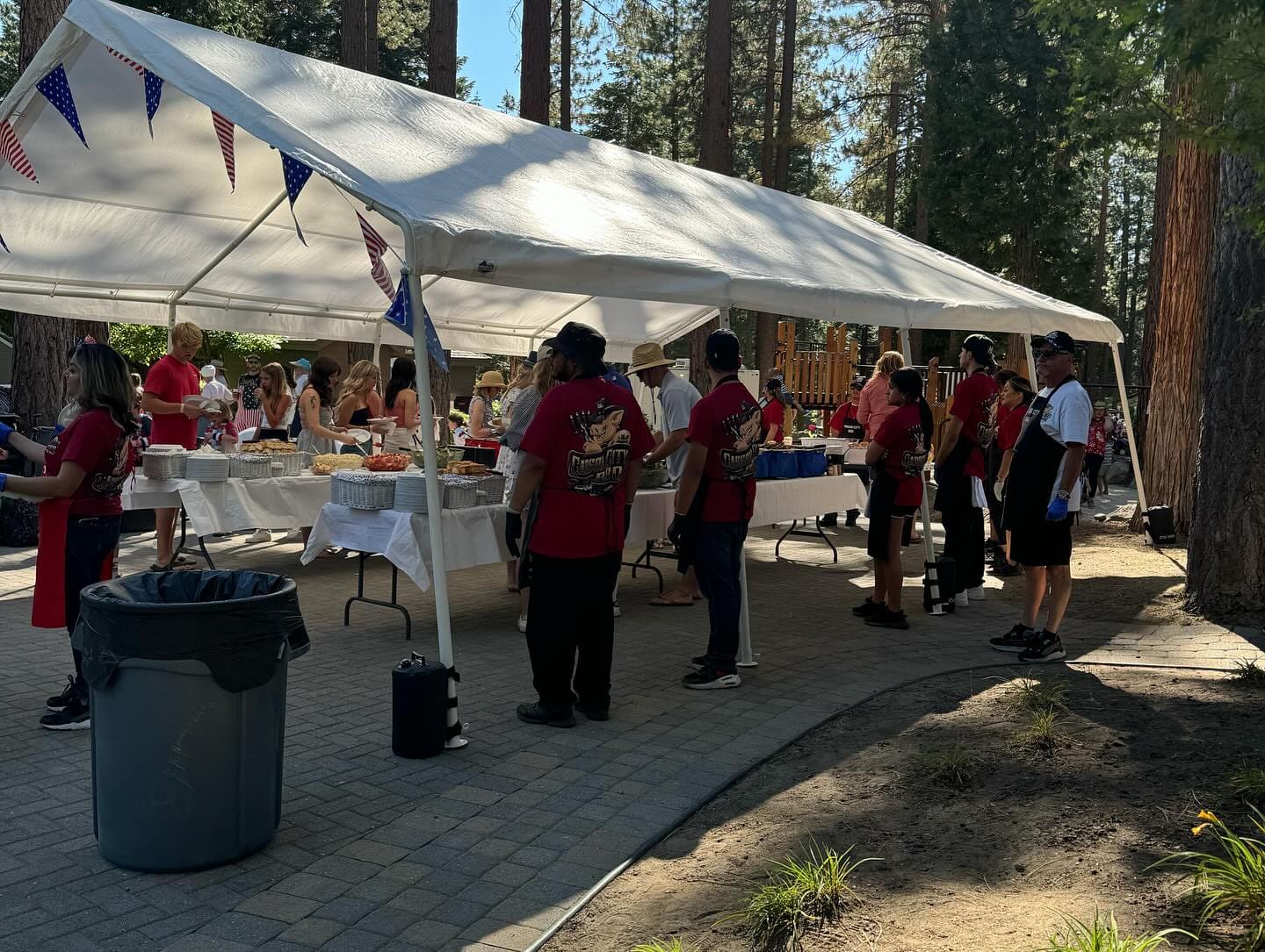 A crew in red Carson City BBQ shirts working a long buffet line beneath an open white marquee strung with bunting, its legs held down by weight bags.
