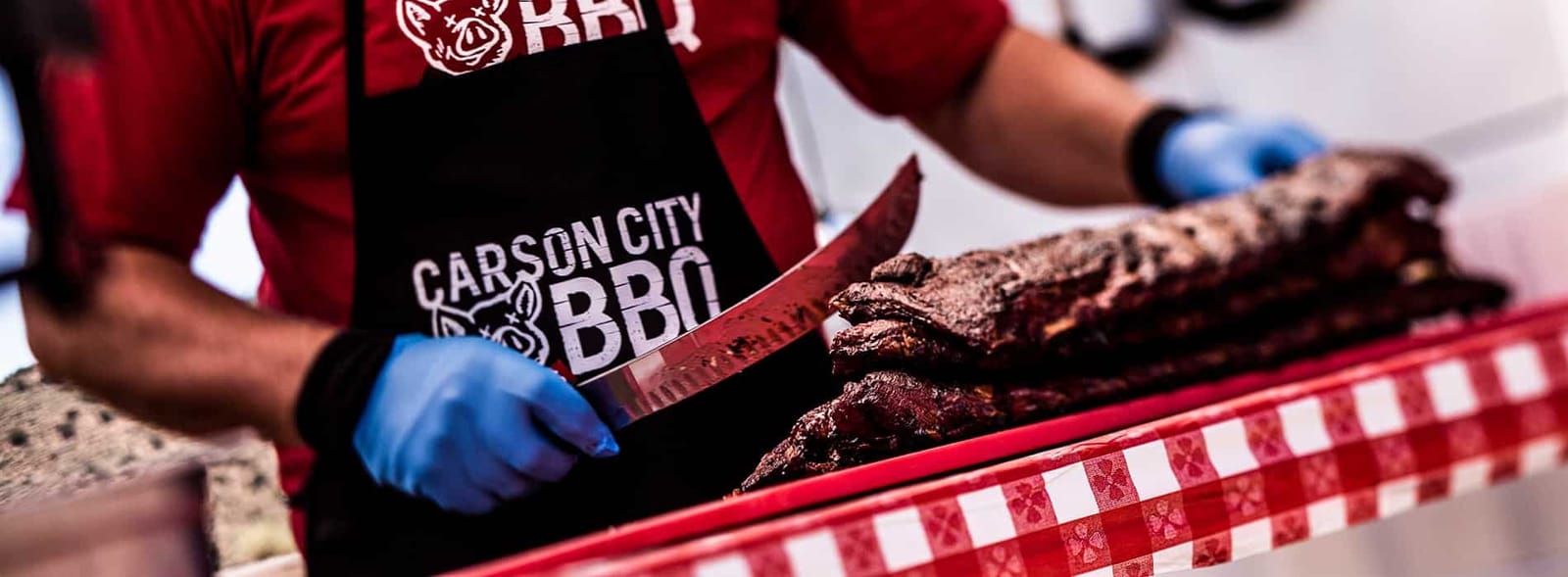 A cook in a Carson City BBQ apron slicing into a rack of smoked ribs on a red-and-white checked cloth