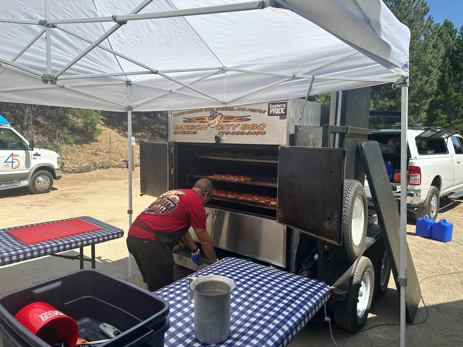 A Carson City BBQ crew member working at the open Southern Pride rotisserie on its trailer, racks of chicken cooking inside, set up under a canopy beside the serving tables.
