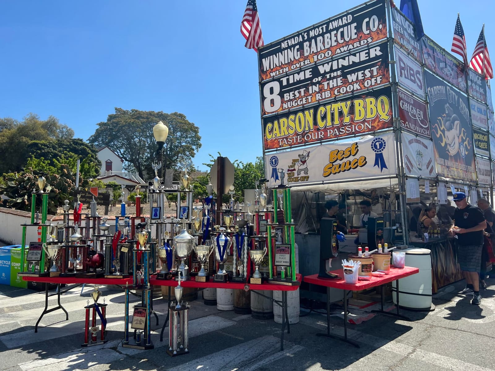 The Carson City BBQ booth on a sunny street, tables of trophies out front beneath a tall sign reading "8 time winner of the Best in the West Nugget Rib Cook-Off".