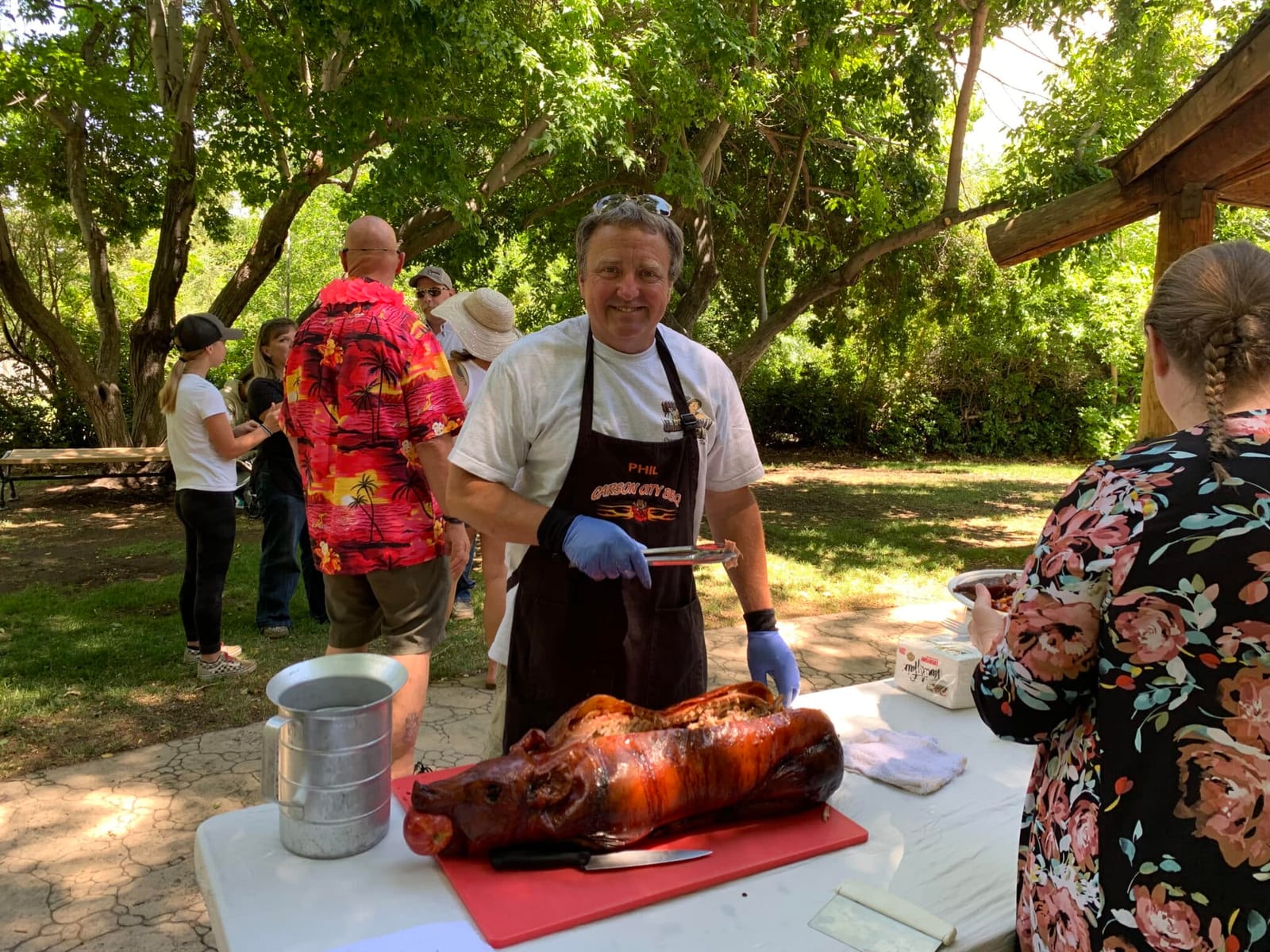 A cook in a Carson City BBQ apron standing behind a whole roasted hog on a red cutting board at a shaded table, with guests gathered on the grass behind him.