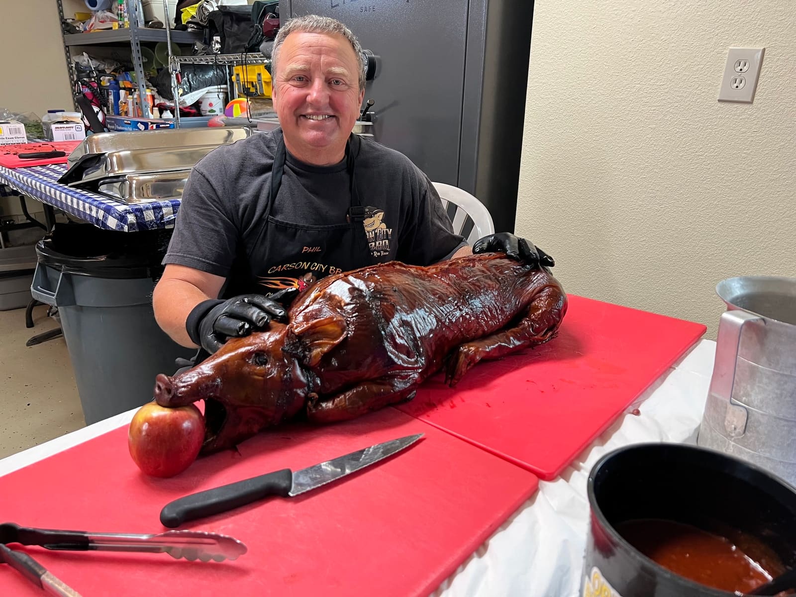 A cook in a Carson City BBQ apron smiling behind a whole roasted suckling pig on a red cutting board, carving knife and tongs laid out beside it.