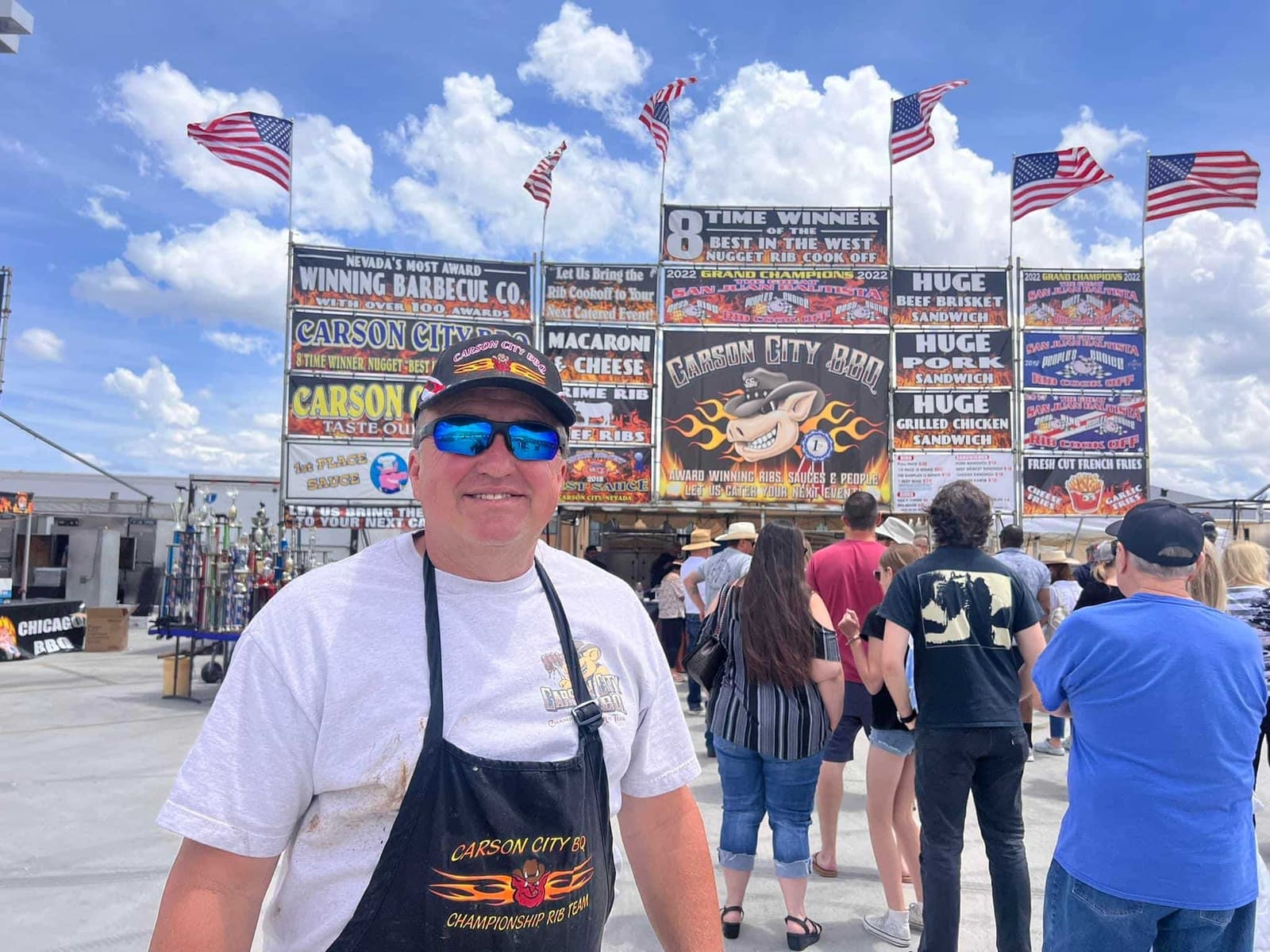 A crew member in a Carson City BBQ apron smiling for the camera in front of the booth's sign board, which is topped by a banner reading "8 time winner, Best in the West Nugget Rib Cook-Off".