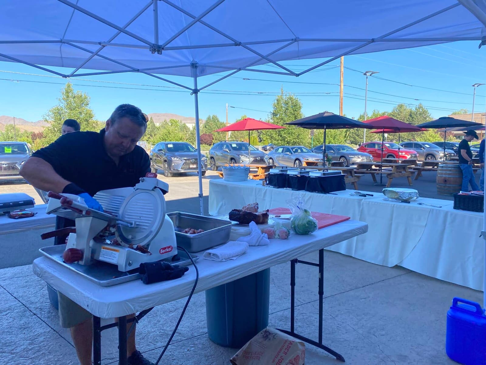 A crew member working a countertop meat slicer at a canopy-shaded serving station in a paved lot, with parked cars, picnic tables under umbrellas and dry hills in the distance.