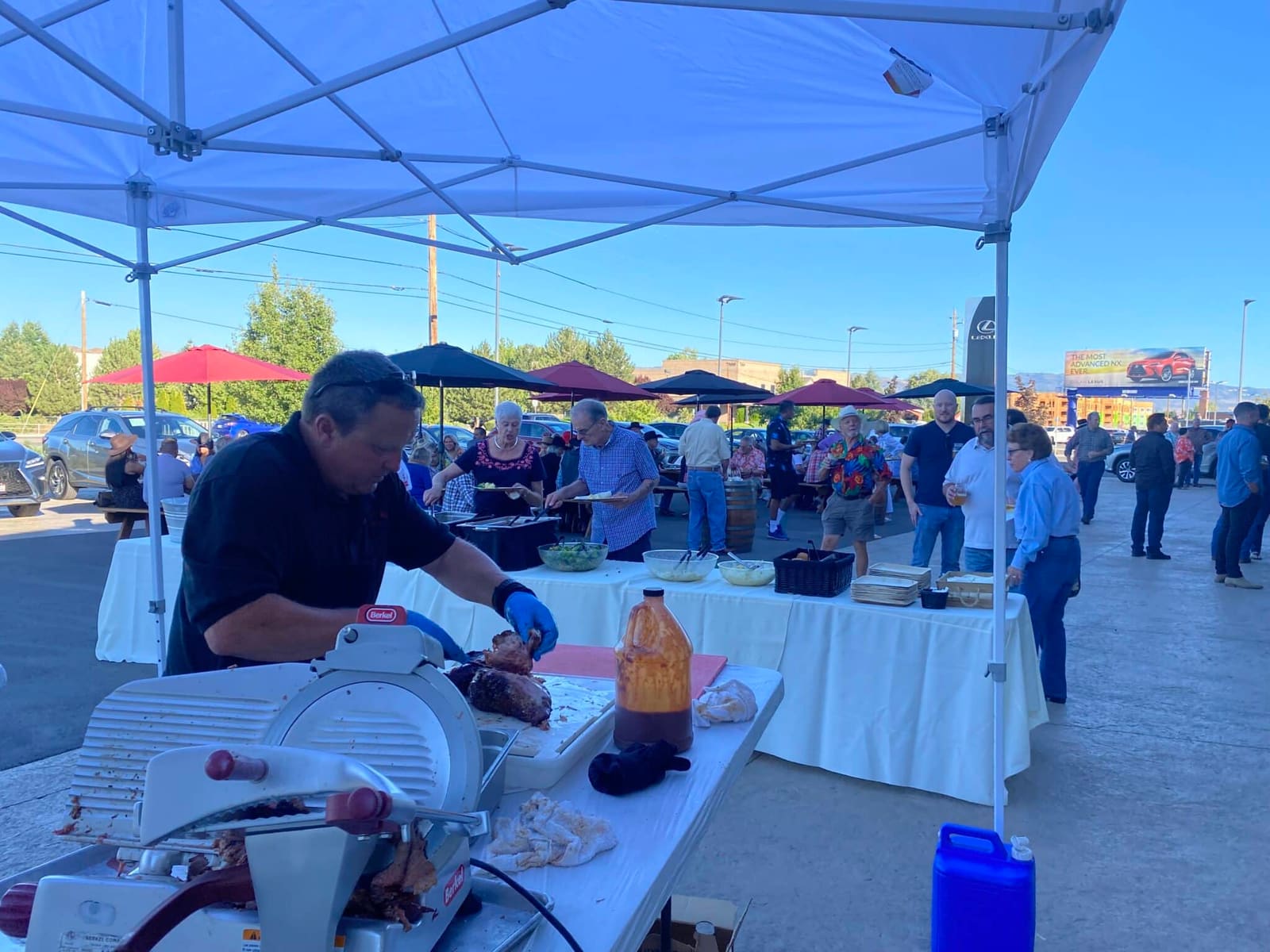 A crew member slicing smoked meat at a canopy-shaded prep table in a paved lot, guests behind him filling plates from a white-clothed buffet beside picnic tables and umbrellas.