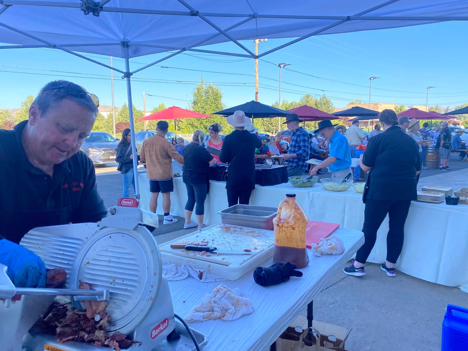 A crew member in a branded shirt working a meat slicer at an outdoor prep table under a pop-up canopy, guests filing along the serving line behind him and office buildings beyond.