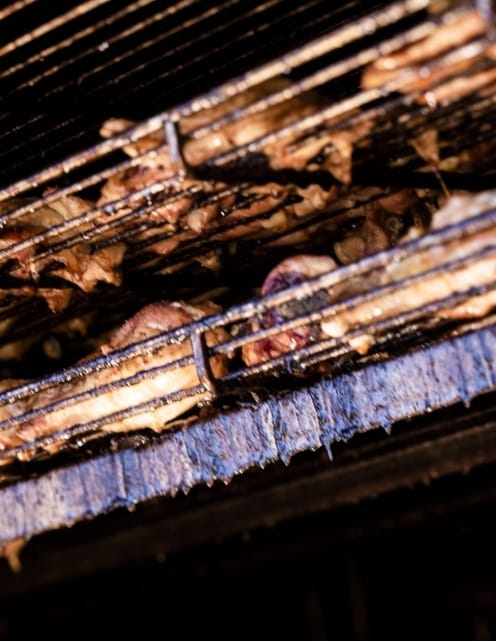 Racks of ribs suspended on the rotisserie shelves of the cooker, lit from below by the fire.