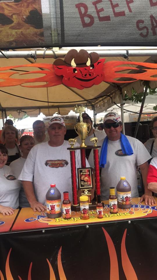 Crew in white shirts and caps standing behind a flame-printed table lined with trophies and bottles of Carson City BBQ sauce at a competition booth.