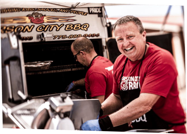 Phil Hyatt in a red Carson City BBQ t-shirt, grinning over his shoulder as he works at the open door of the Carson City BBQ cooker trailer, another crew member tending the racks behind him.