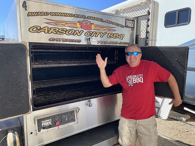 Phil Hyatt in a red Carson City BBQ shirt and sunglasses standing with one arm raised beside the open doors of the Southern Pride cooker trailer.