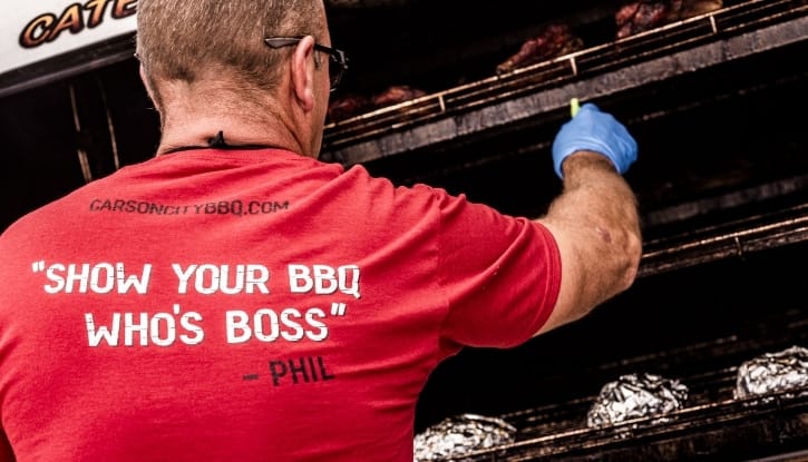 The back of a red Carson City BBQ shirt reading “Show your BBQ who's boss” — Phil, worn by a cook in a blue glove reaching into the open cooker to move racks of meat.
