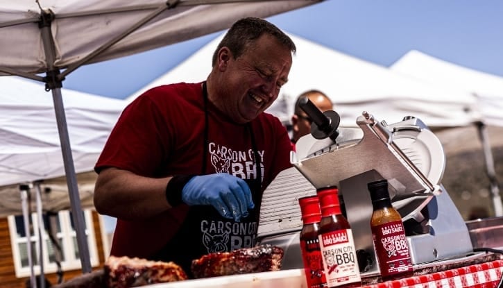 Phil Hyatt working a meat slicer at an outdoor catering station under a canopy, two bottles of Carson City BBQ sauce and a tray of ribs on the table beside him.