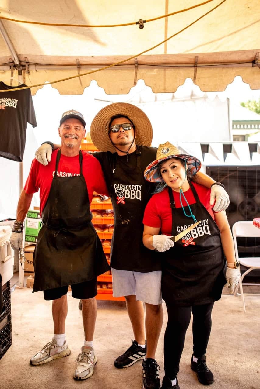 Three Carson City BBQ crew members posing together in their aprons under the tent at a cook-off, laughing with their arms around each other.