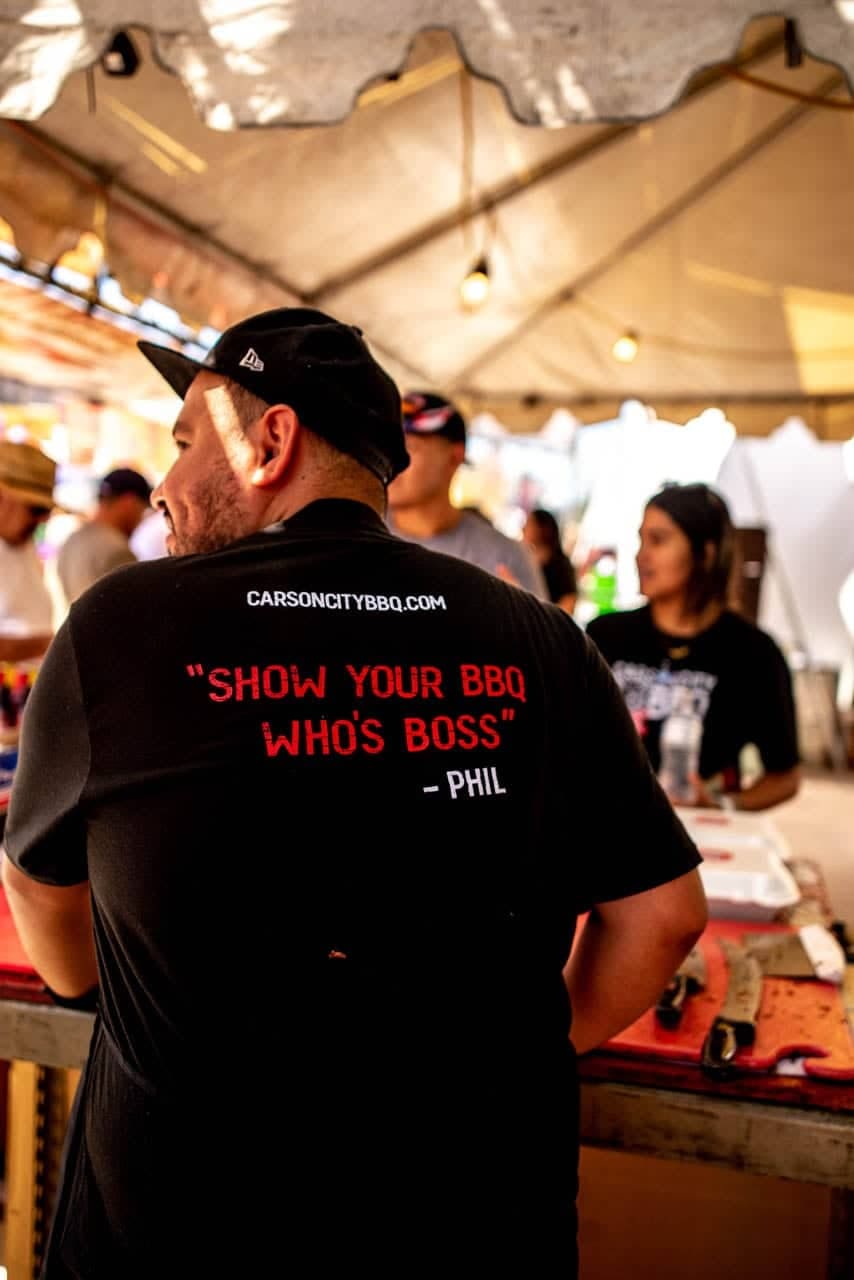 The back of a crew member's black Carson City BBQ shirt, printed with the slogan "Show your BBQ who's boss" credited to Phil.