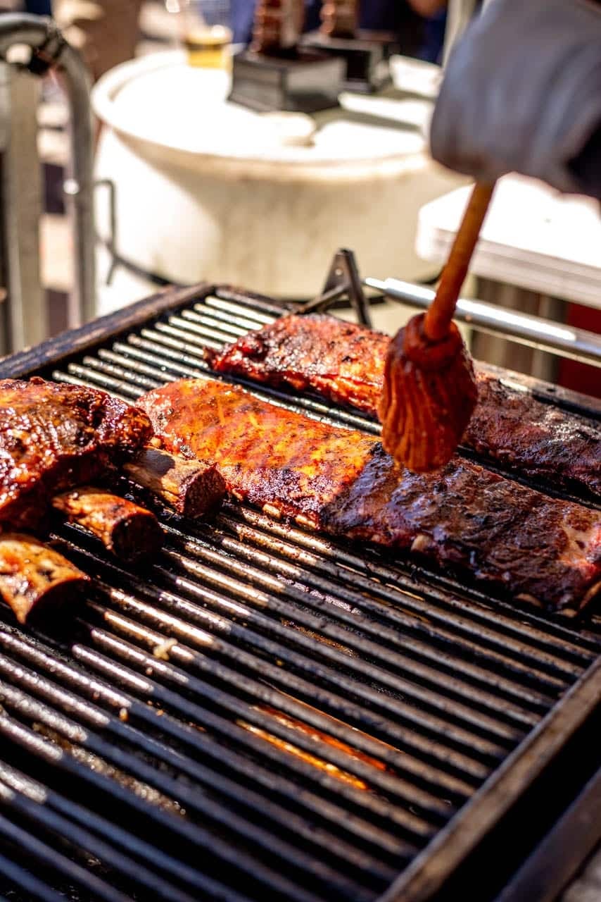 A basting brush painting sauce onto racks of ribs laid across a black grill grate.