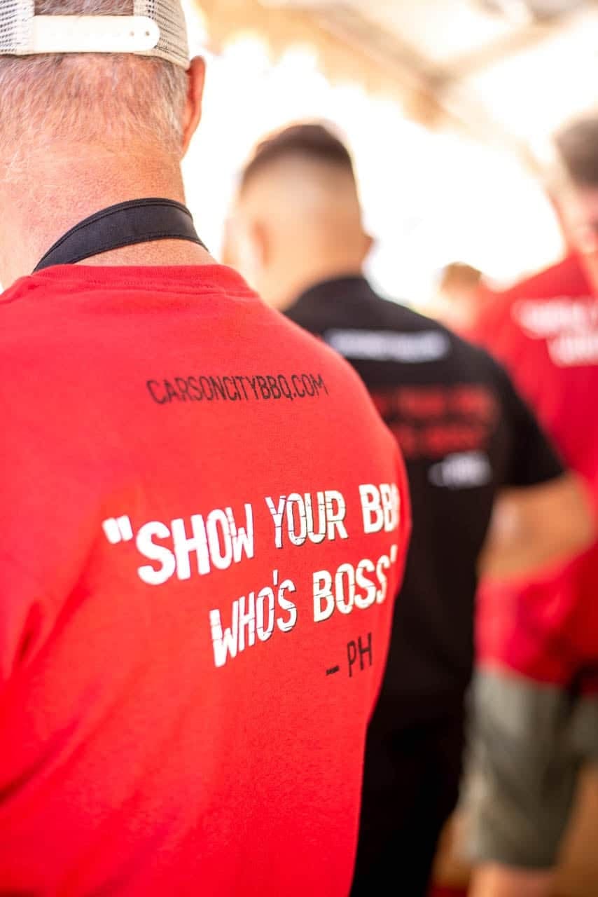 The back of a red Carson City BBQ shirt reading "Show your BBQ who's boss", worn by a crew member working the line at a cook-off.