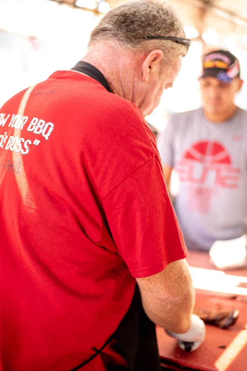 A Carson City BBQ crew member in a red shirt working head-down at the prep table under the tent, a colleague behind him.
